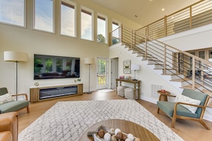 A spacious living room with a large flat-screen TV, a fireplace, a staircase, and a high ceiling with large windows. - Hot Tub, Dock, Game Room! Stunning Lake Wylie Home (Charlotte)
