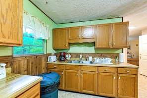 A kitchen with wooden cabinets, a sink, a window with curtains, and a trash can.