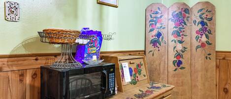 A microwave on a wooden counter with a basket, a bag of chips, and a framed picture.