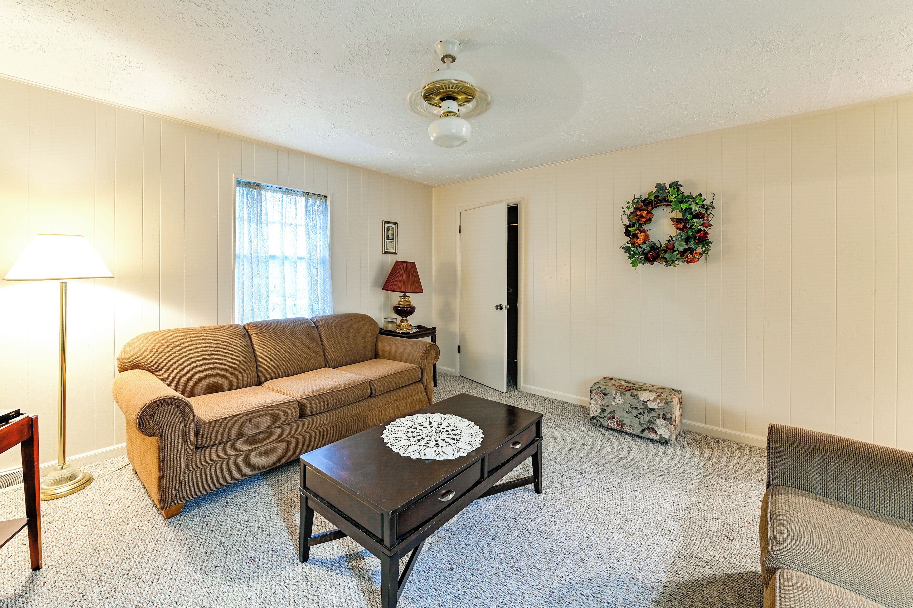 A living room with a brown sofa, a dark wooden coffee table with a doily, a floor lamp, a side table with a lamp, a door, and a wreath on the wall.