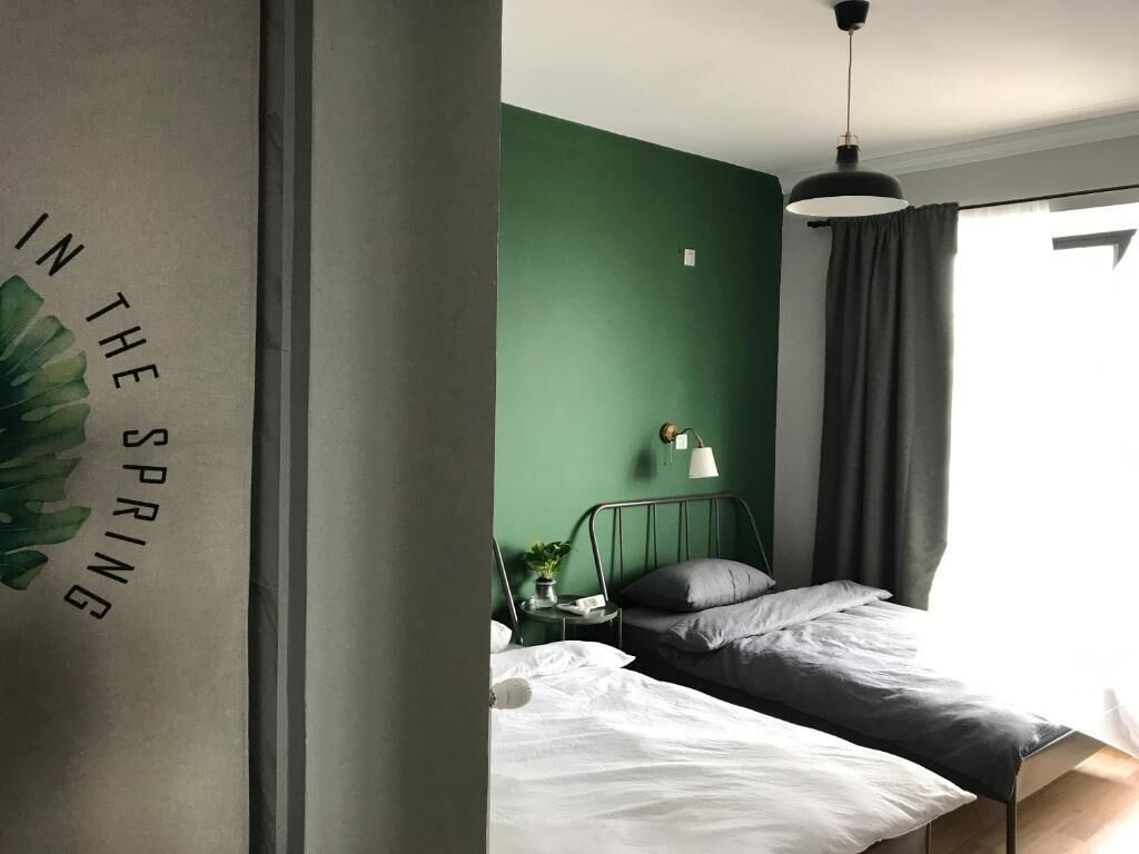 Un dormitorio con pared verde, una cama con ropa de cama blanca y gris, una mesita de noche con una lámpara y una ventana con cortinas.
