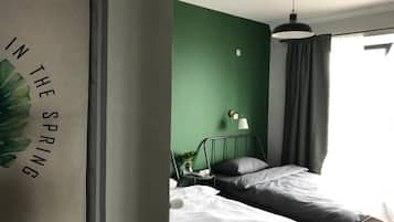 Un dormitorio con pared verde, una cama con ropa de cama blanca y gris, una mesita de noche con una lámpara y una ventana con cortinas.