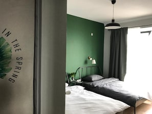 A bedroom with a green wall, a bed with white and grey bedding, a nightstand with a lamp, and a window with curtains. - Ma Cherie Hostel (Shanghai)