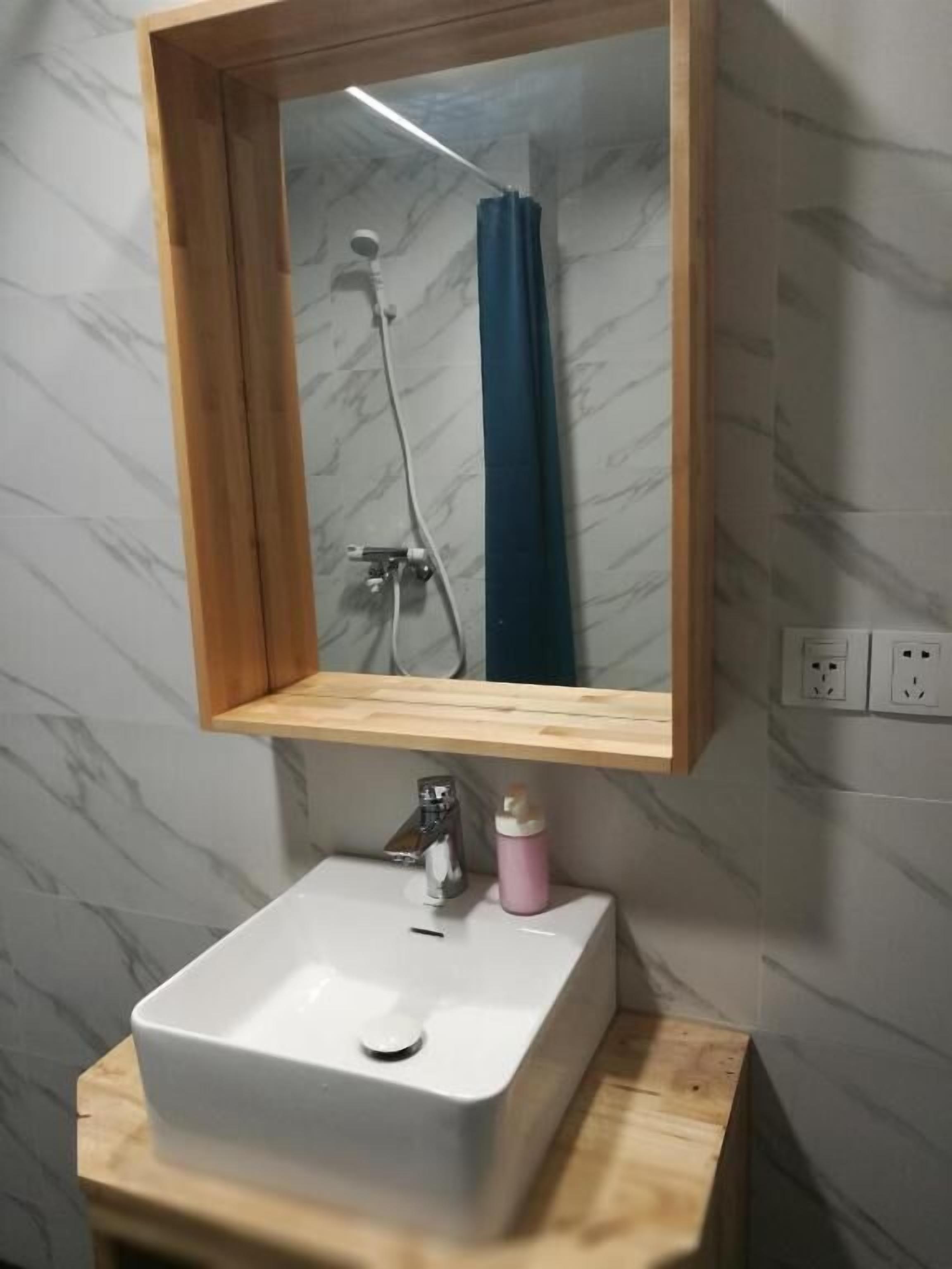 A bathroom with a marble wall, a wooden-framed mirror, a white sink, and a shower visible in the mirror.