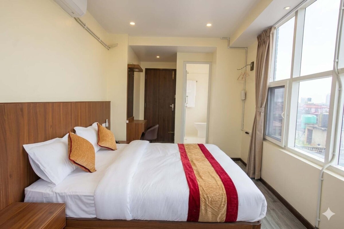 Habitación de hotel con cama, cabecera de madera, ventana con vistas y puerta que da a un baño.