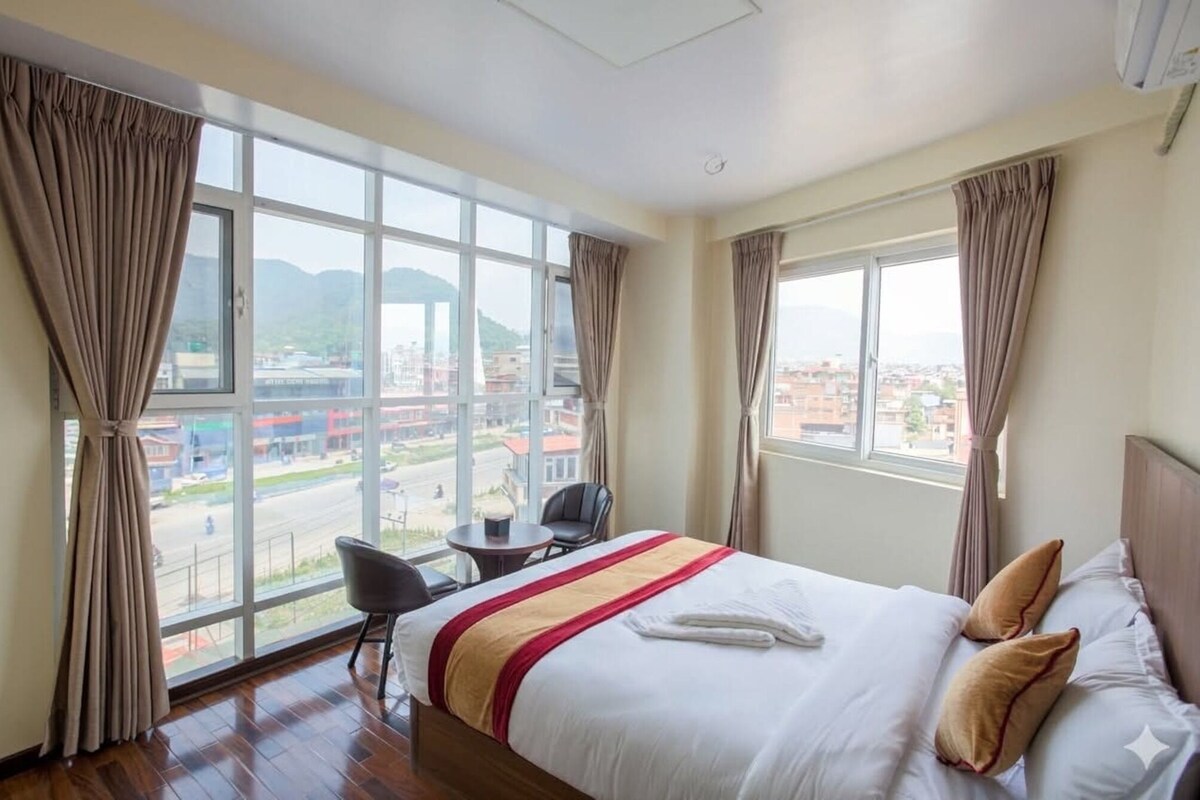 Habitación de hotel con cama, una silla, una mesita y ventanas amplias con vistas a la ciudad.
