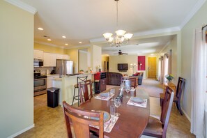 A dining area with a wooden table set for six, surrounded by chairs, with a kitchen and living area in the background.