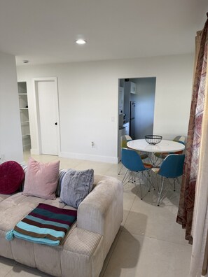 A living room with a sofa, a dining table with chairs, a kitchen area, and a door.