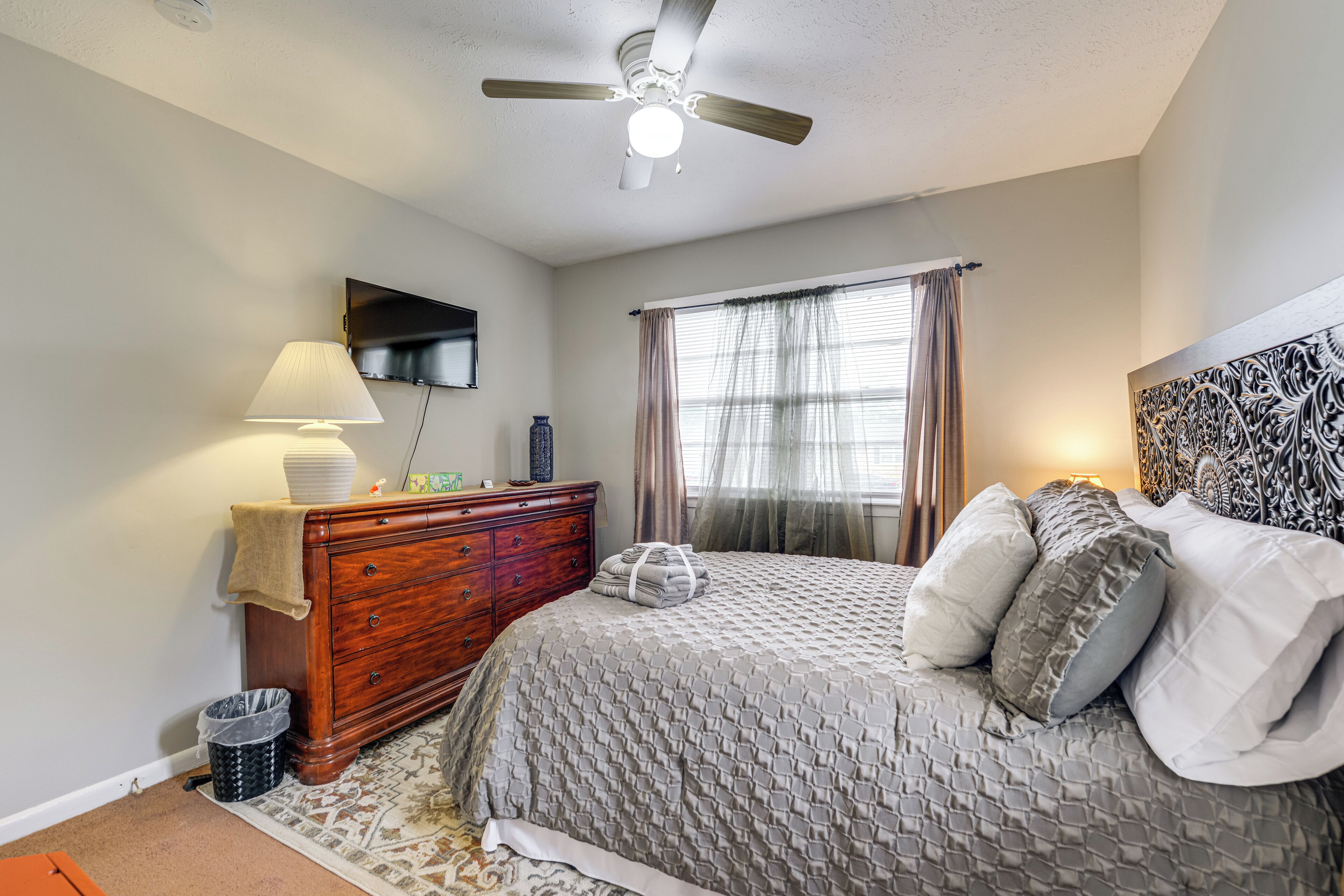 A bedroom with a bed, a ceiling fan, a dresser, a TV, and a window with curtains.