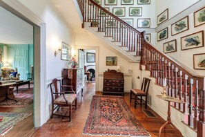 A hallway with a wooden staircase, a dining area with chairs and a table, and a patterned rug on the floor. - Private, Exclusive, Historic Pocomoke River Estate (Pocomoke City)