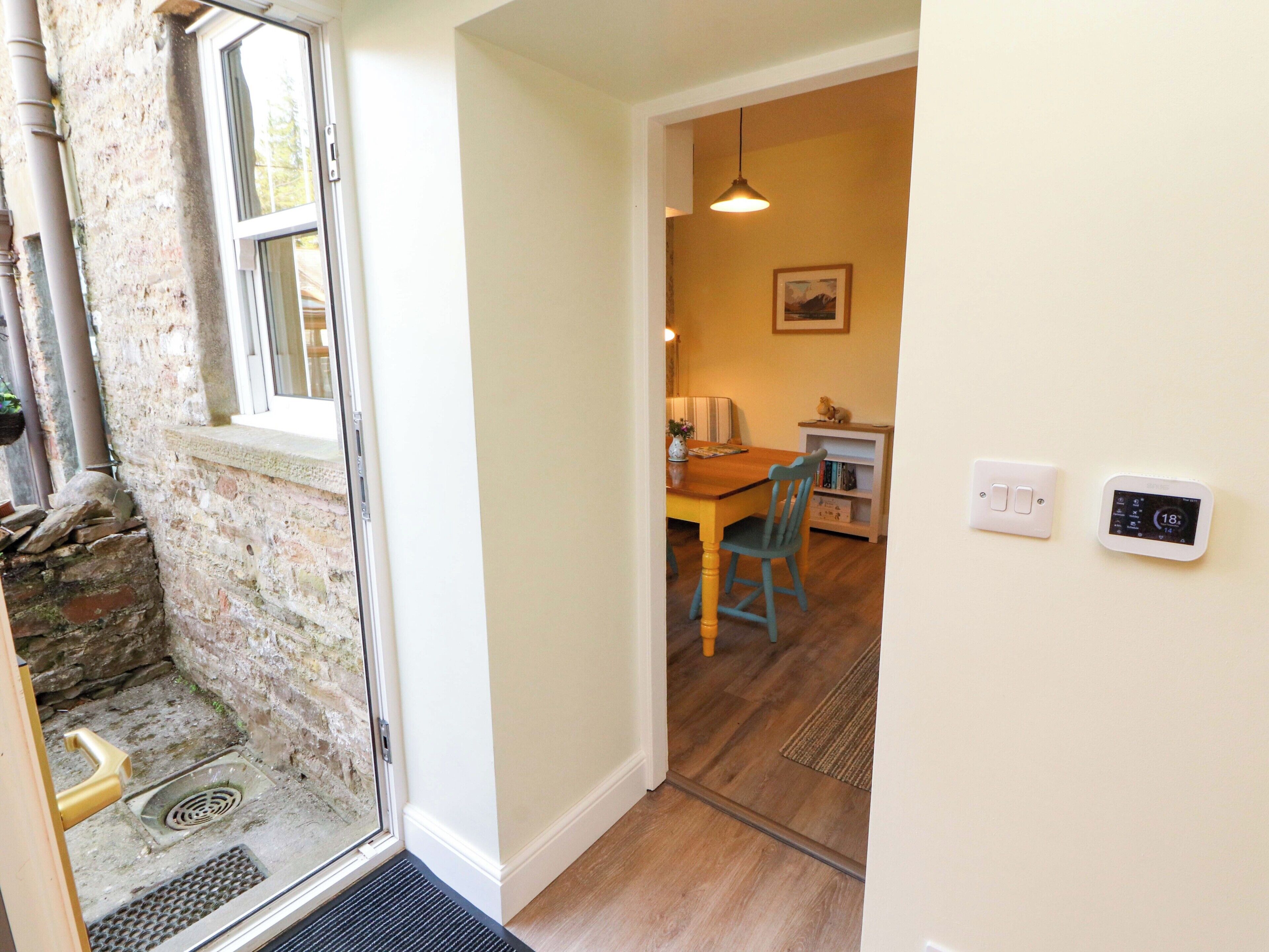 A room with a stone wall, a wooden door, a window, a dining table with chairs, a wall-mounted thermostat, and a light fixture.