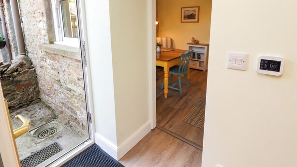 A room with a stone wall, a wooden door, a window, a dining table with chairs, a wall-mounted thermostat, and a light fixture. - East View (Kirkby Stephen)