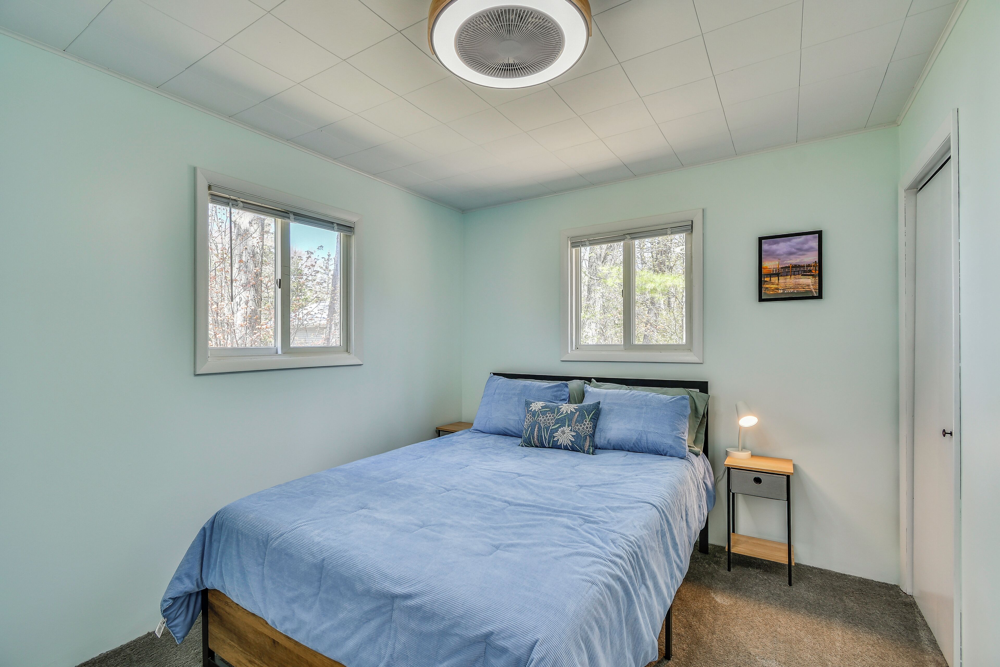 A bedroom with a bed, two windows, a nightstand with a lamp, and a door.