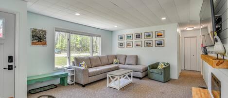 A living room with a sectional sofa, a coffee table, a bench, and a dining table. There are framed pictures on the wall and a window with a view of trees.
