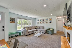 A living room with a sectional sofa, a coffee table, a bench, and a dining table. There are framed pictures on the wall and a window with a view of trees.