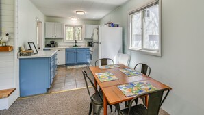 Una cocina con armarios blancos, un refrigerador, una mesa de comedor con sillas y una ventana con persianas.