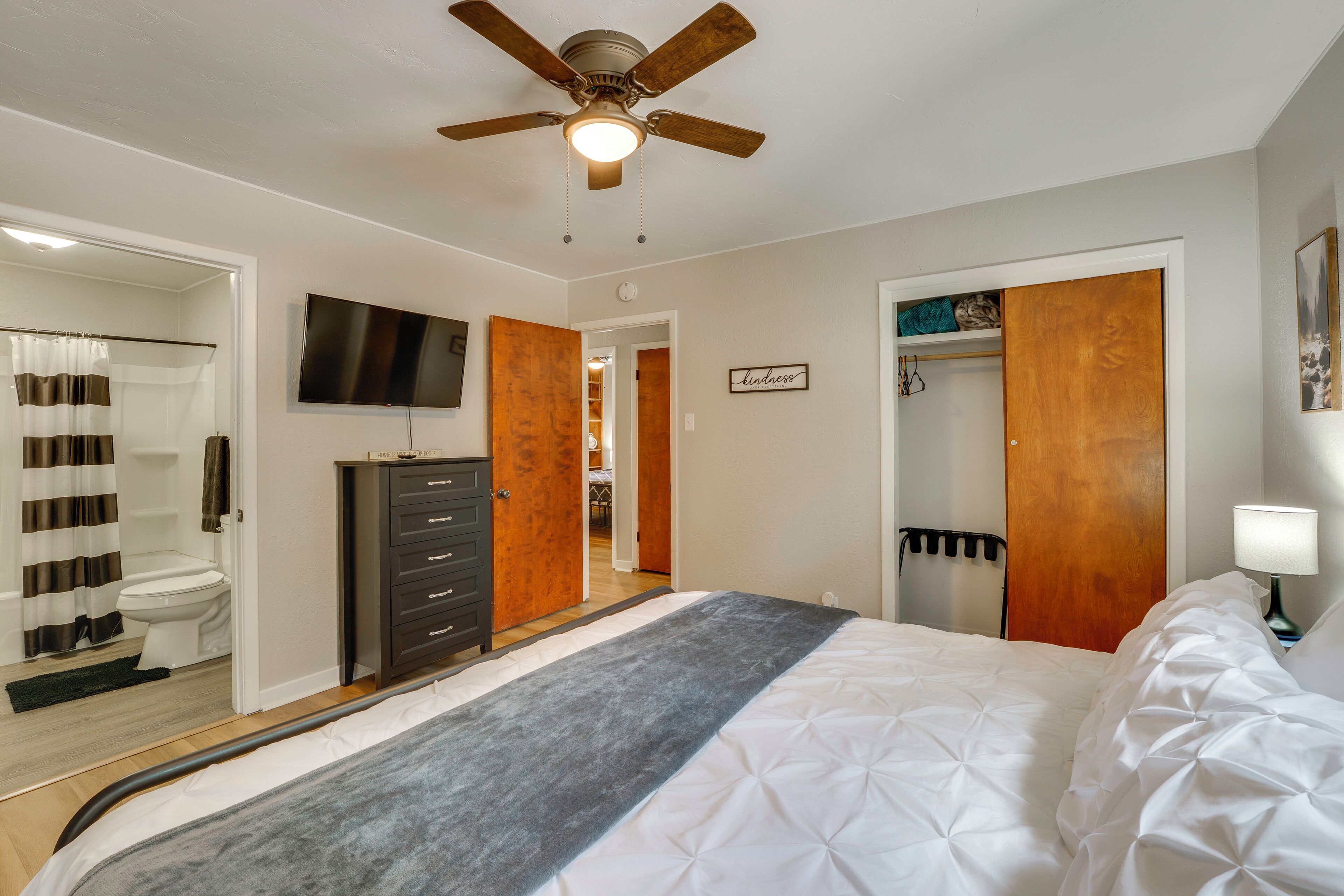 Une chambre à coucher comprenant un lit, un ventilateur de plafond, une télévision, une commode, un placard et une salle de bain avec des toilettes et un rideau de douche.