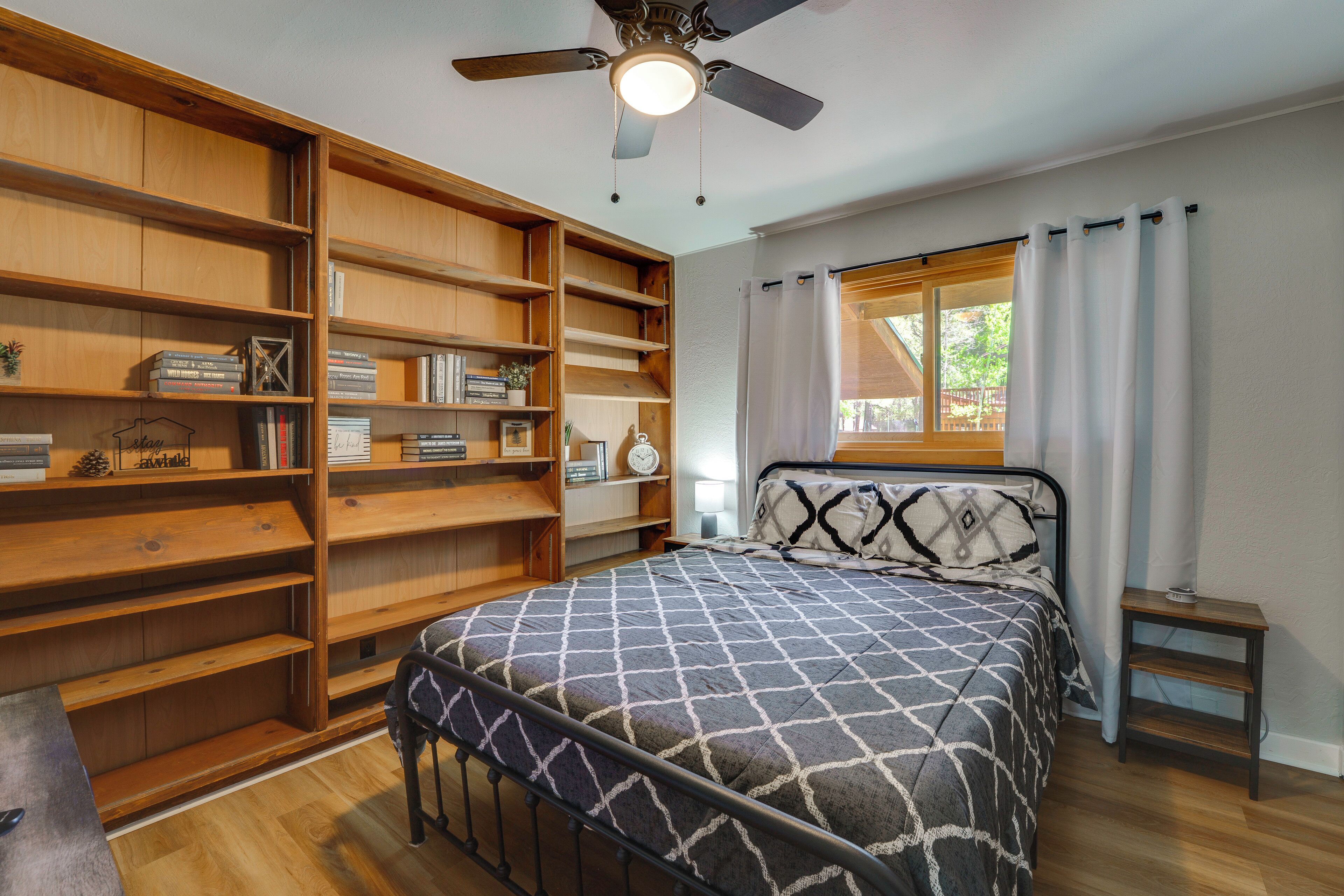 Un dormitorio con cama, ventilador de techo, estanterías de madera, ventana con cortinas y una pequeña mesa de madera.