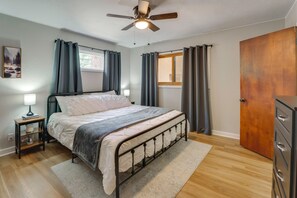 Ein Schlafzimmer mit Bett, Nachttisch, Deckenventilator, Fenster mit Vorhängen, Holztür und Kommode.