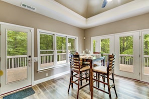 A dining area with a table and chairs, large windows with a view of trees, and a ceiling vent.