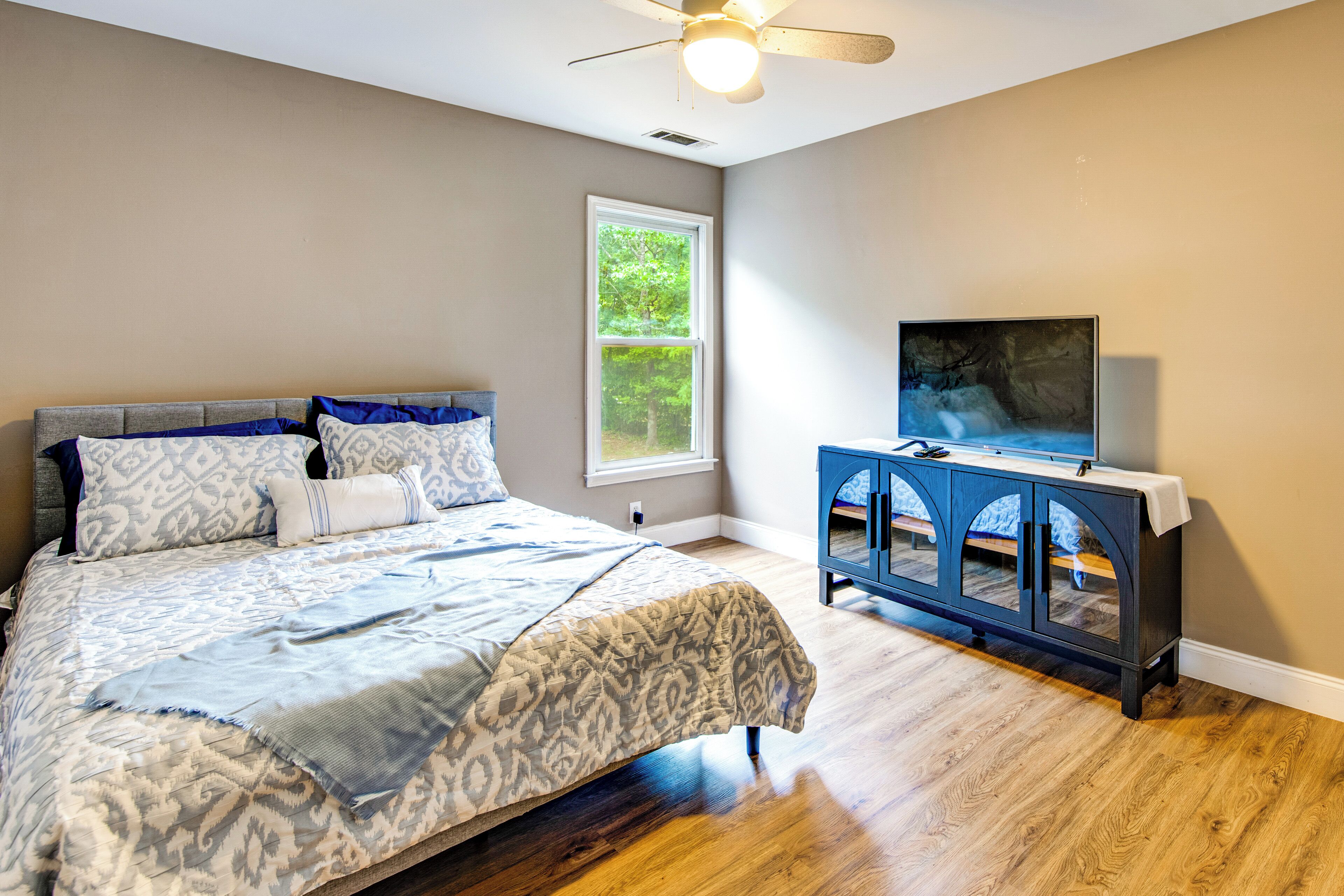 A bedroom with a bed, a window, a ceiling fan, and a TV on a stand.