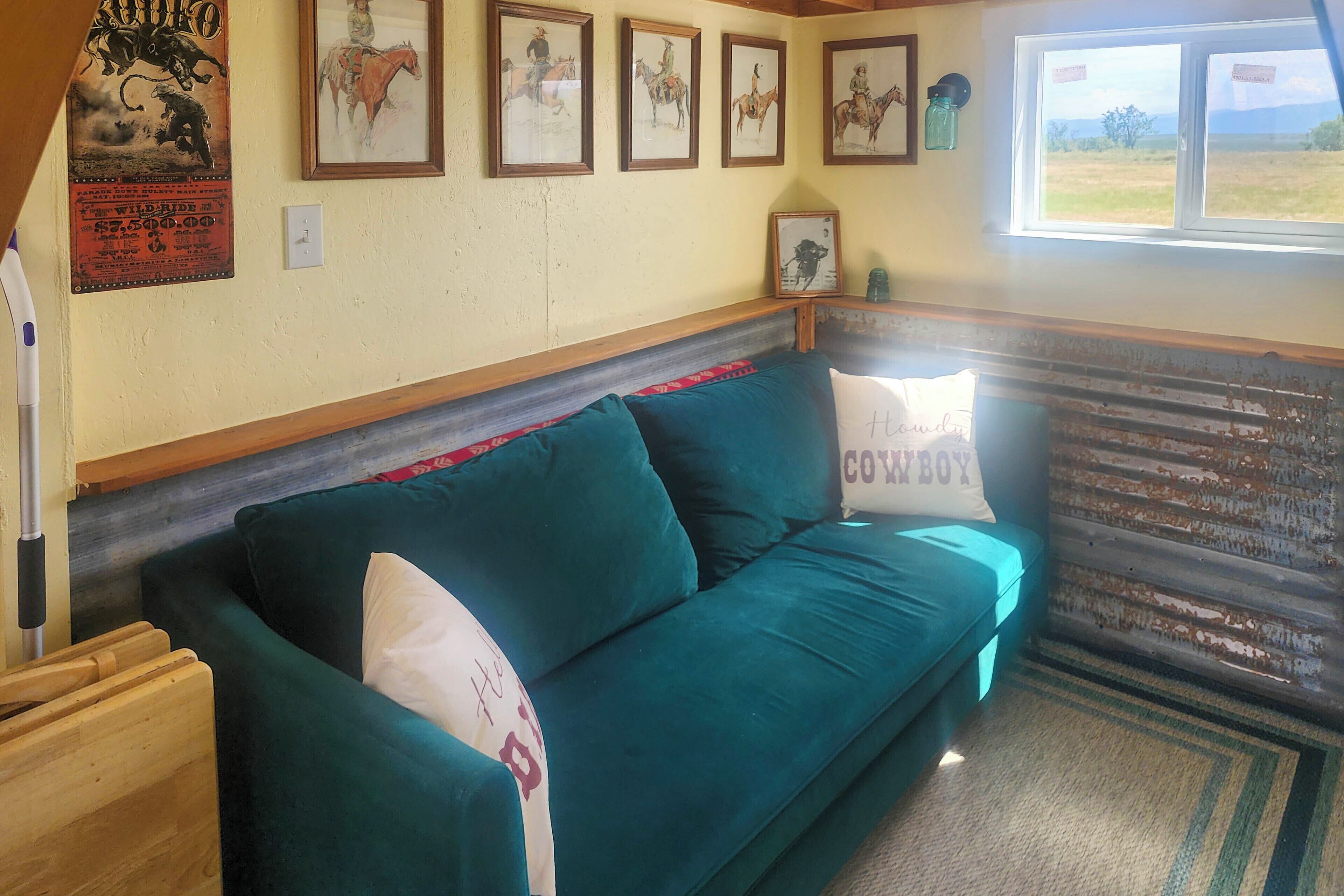 A teal sofa with pillows, a wooden headboard, framed cowboy-themed artwork, and a window with a view of the outdoors.