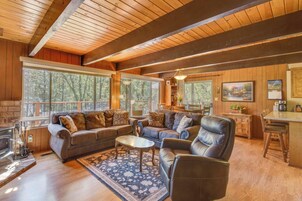 A cozy cabin interior with a fireplace, wooden walls, large windows, a dining area, and a kitchen.