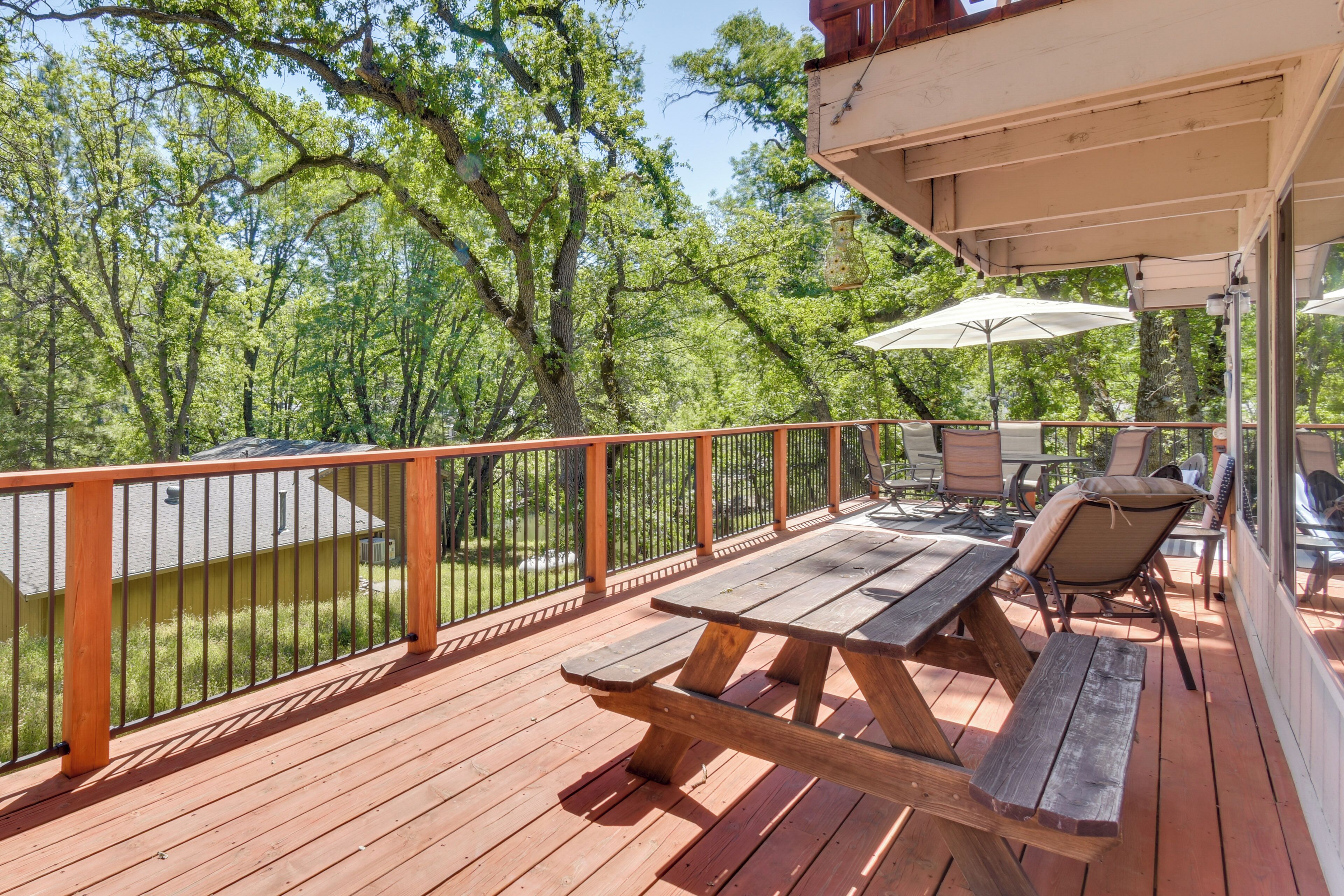 A wooden deck with picnic tables and chairs, surrounded by a metal railing, overlooking a wooded area.