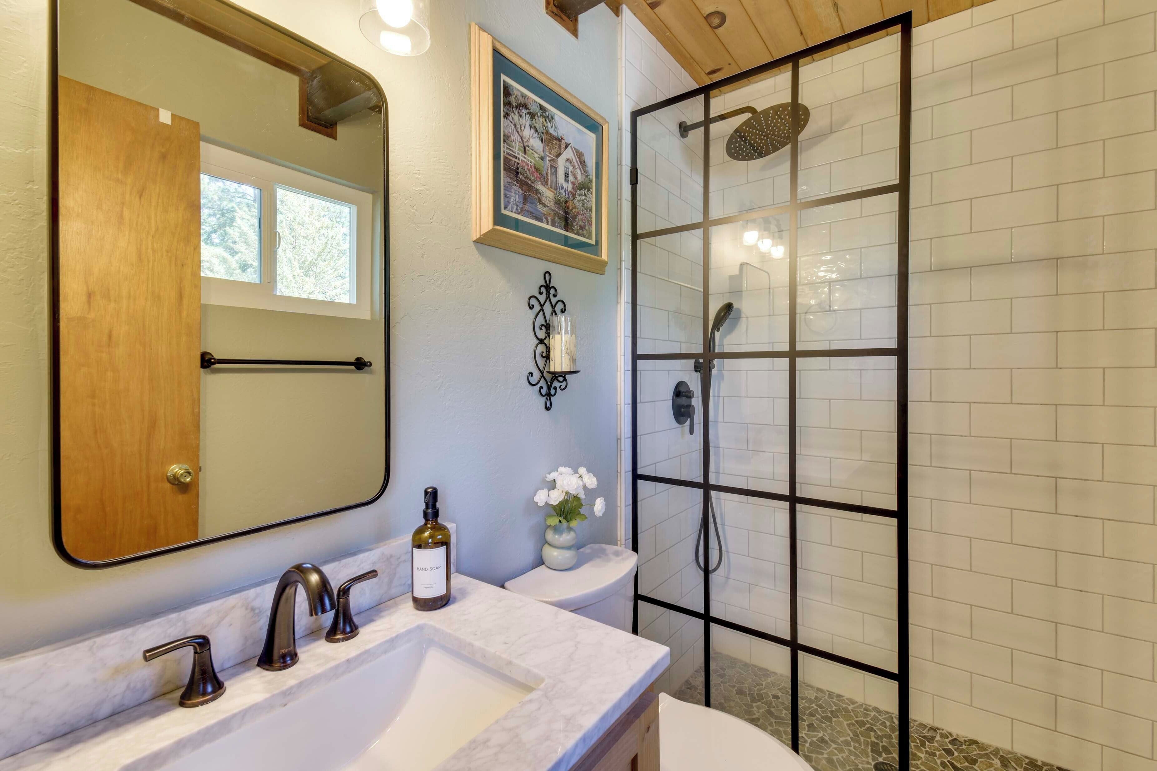 A bathroom with a marble sink, a wooden door, a framed picture, a vase with flowers, and a glass-enclosed shower.