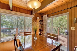 A wooden-paneled room with a dining table, chairs, a vase with plants, and large windows offering a view of trees.