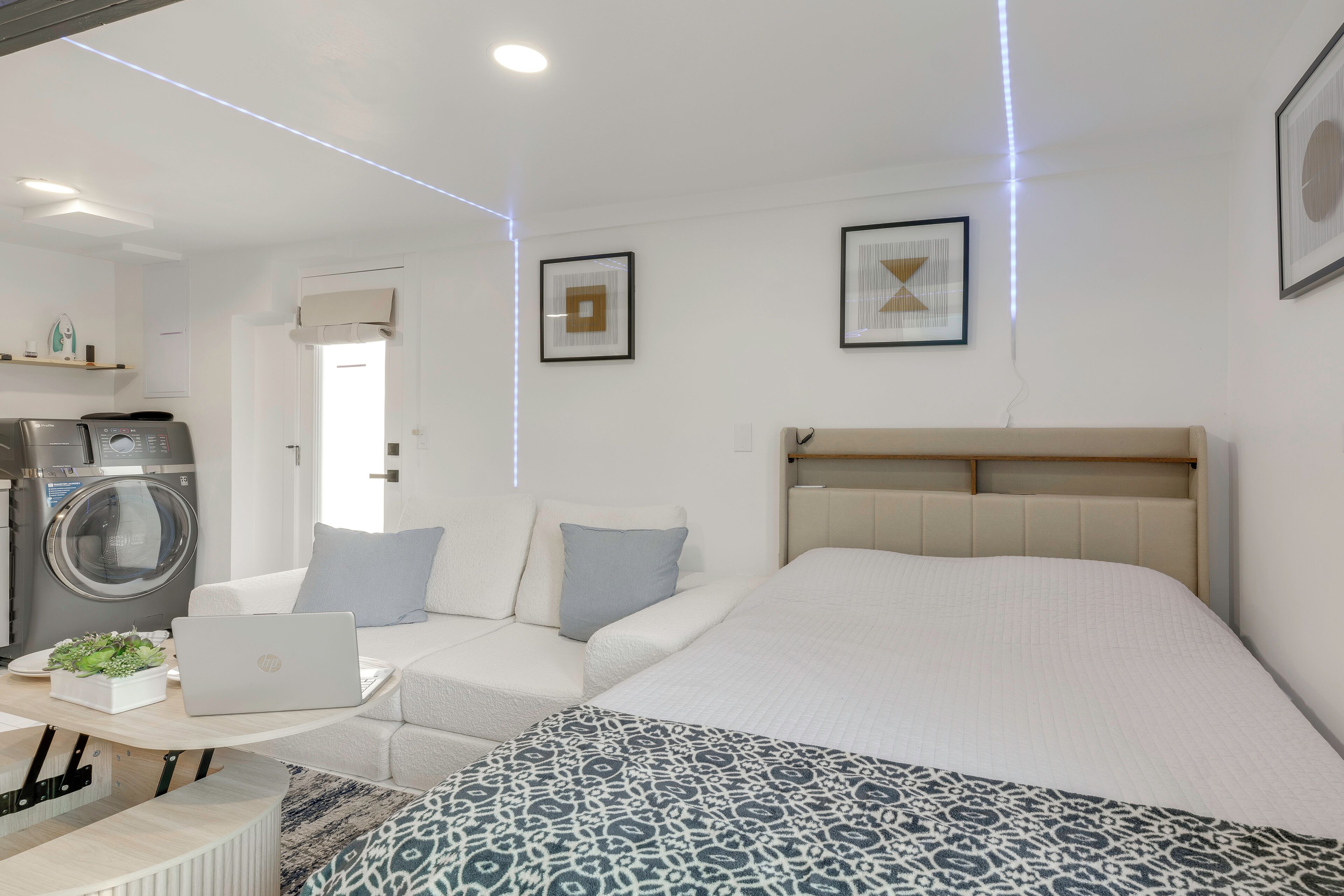 A modern hotel room with a bed, a sofa, a table with a laptop, a washing machine, and framed artwork on the walls.