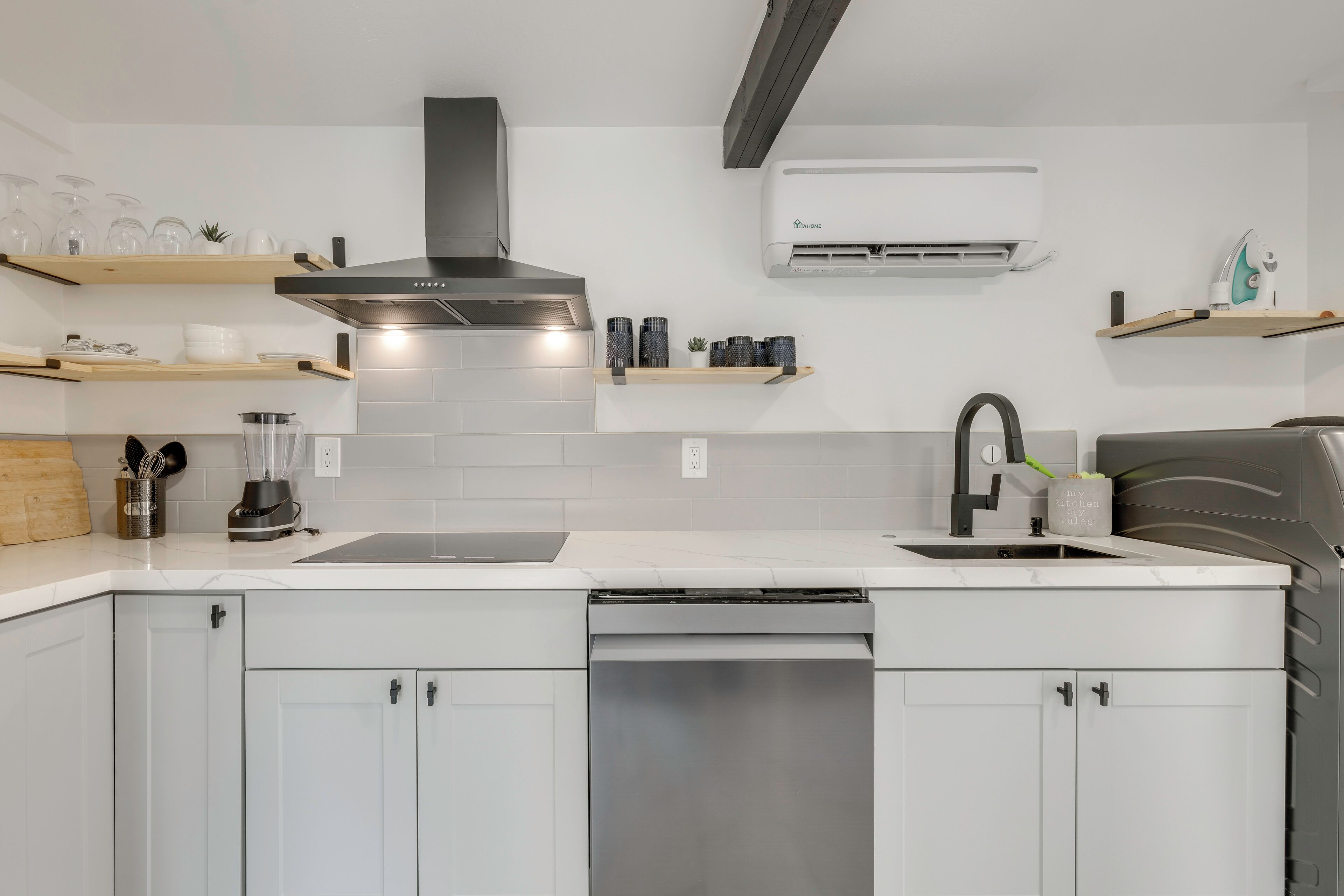 Una cocina moderna con armarios blancos, lavavajillas de acero inoxidable, campana extractora negra y aire acondicionado de pared.