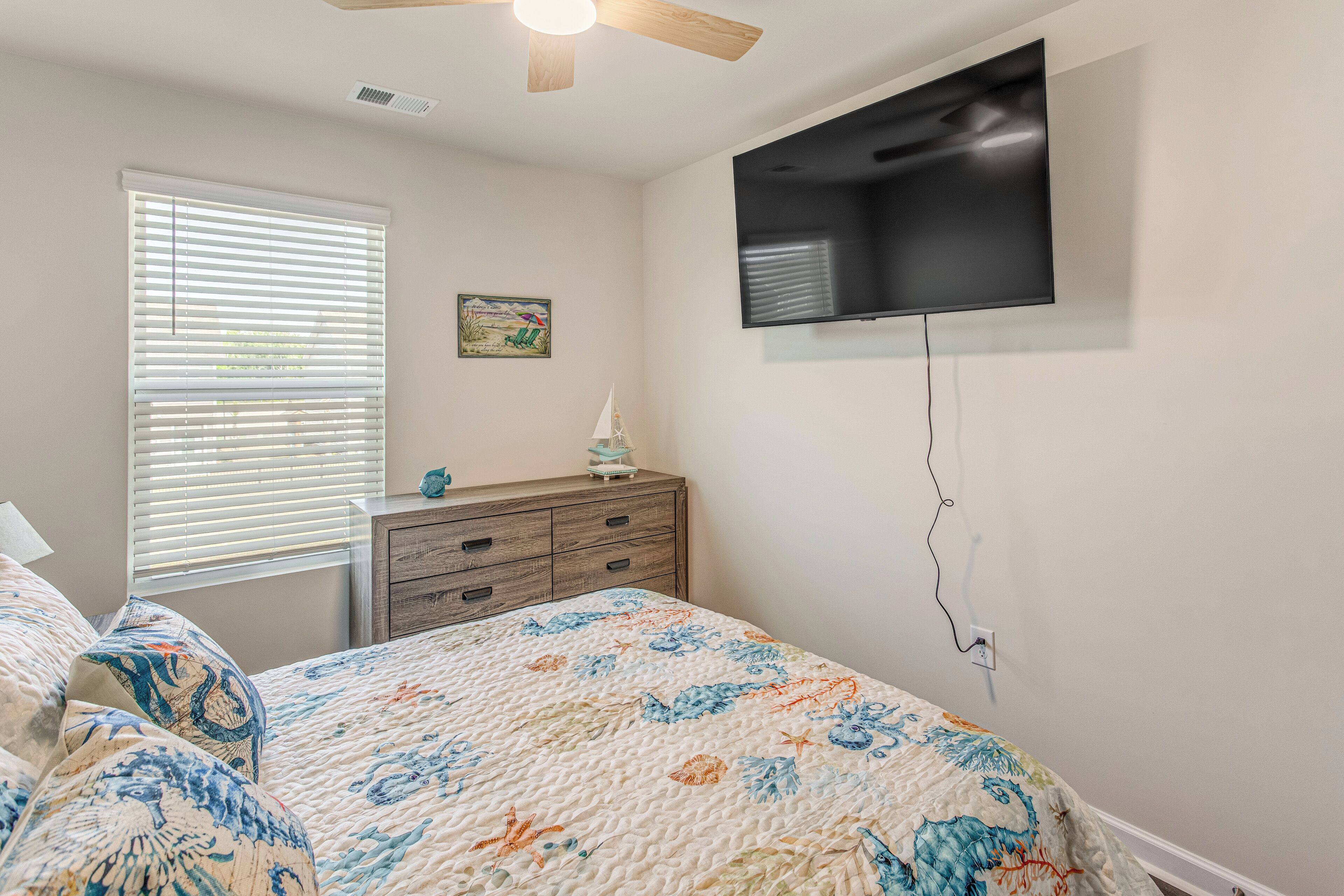 Quarto com cama, cômoda, televisão fixada na parede, ventilador de teto e janela com persianas.