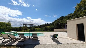 Piscina con sillones de descanso y una sombrilla, rodeada de follaje y cielo despejado.