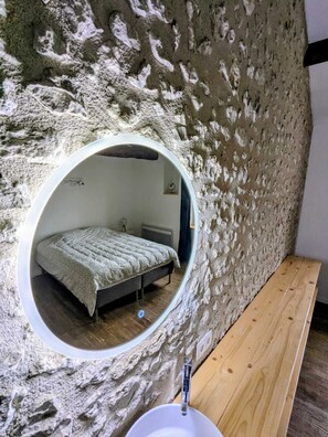 A circular mirror reflecting a bed and a wooden bench in a room with textured walls. - Charming holiday house with stunning view and heated swimming pool. (Palluaud)