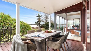 Een overdekt terras met rieten meubels, een tafel met een boek en een fles, en uitzicht op zee en bomen.