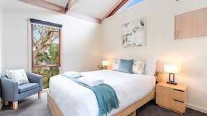 Une chambre à coucher comprenant un lit, une chaise, une table de chevet, une lampe, une fenêtre donnant sur des arbres et un tableau accroché au mur.