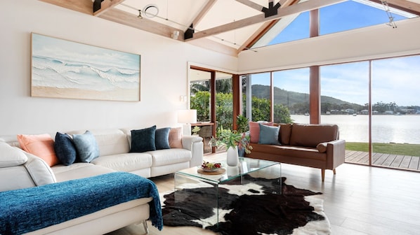 A modern living room with a glass coffee table, a white sofa with blue and peach cushions, a brown sofa with blue cushions, and a large window overlooking a body of water.