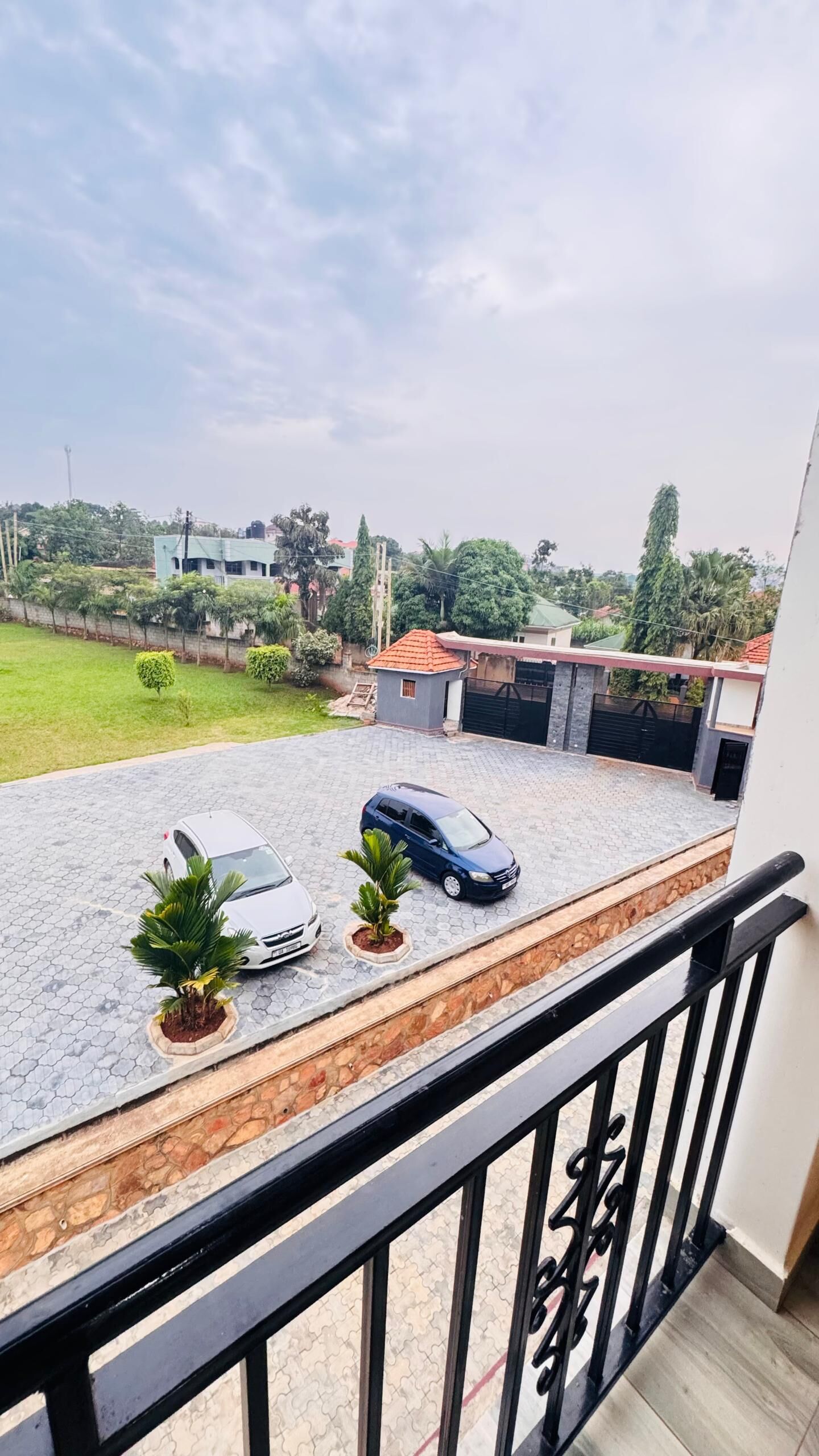 A balcony with a view of a paved driveway, two cars, and a small building with a red roof.