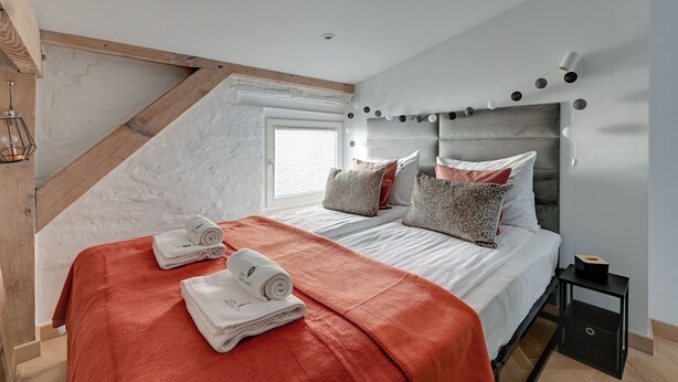 Quarto com teto inclinado, cama com roupa de cama branca, manta vermelha e cabeceira com luzes.