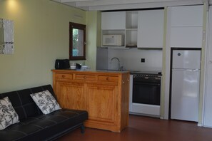Eine kleine Küche mit Einbauofen, Mikrowelle und Kühlschrank. Eine Holzeinrichtung mit Schubladen befindet sich neben der Küche, und ein schwarzes Ledersofa mit floralen Kissen ist im Vordergrund abgebildet.