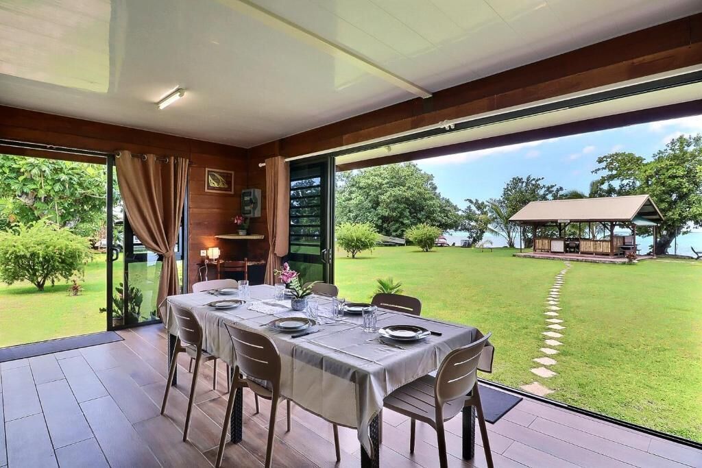 Un espace extérieur aménagé pour les repas, avec une table dressée pour quatre personnes, une porte vitrée donnant sur un jardin et une vue sur une pelouse et un bâtiment situé au loin.