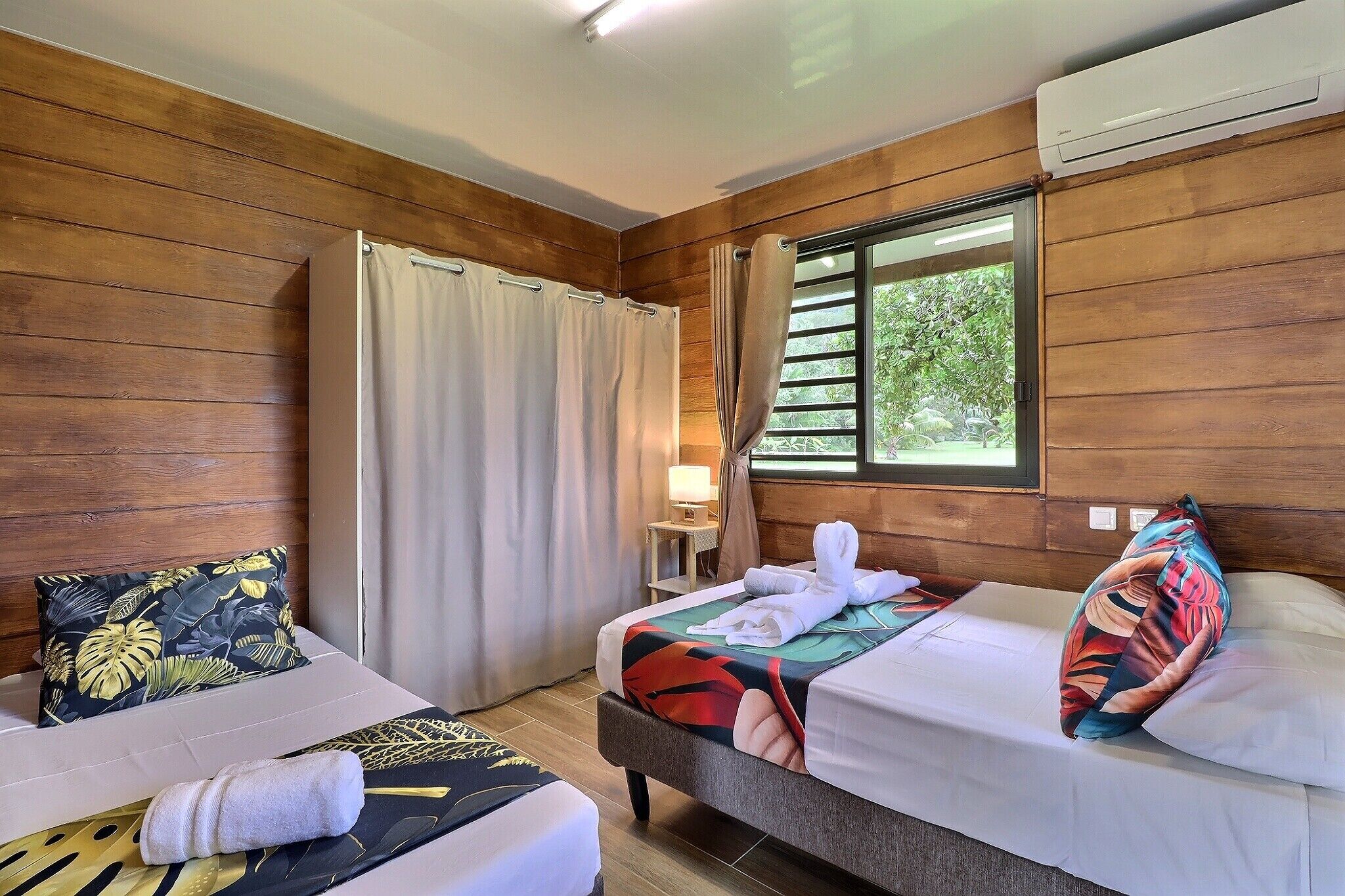 A hotel room with wooden panel walls, a bed with a floral bedspread, a bedside table with a lamp, and a window with a view of greenery.