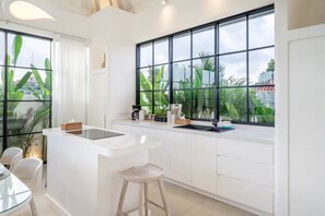 Una cocina moderna con encimera blanca, taburetes de bar, cafetera y grandes ventanales con vistas a zonas verdes.