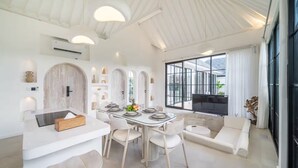 A modern dining area with a glass table, white chairs, and a large window with a view. - White Shell Villa 1 (badung)