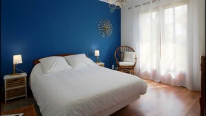 Uma cama bem arrumada com lençóis brancos, uma mesinha de cabeceira de madeira com abajur, um objeto decorativo na parede, uma cadeira de vime e cortinas leves em uma janela.