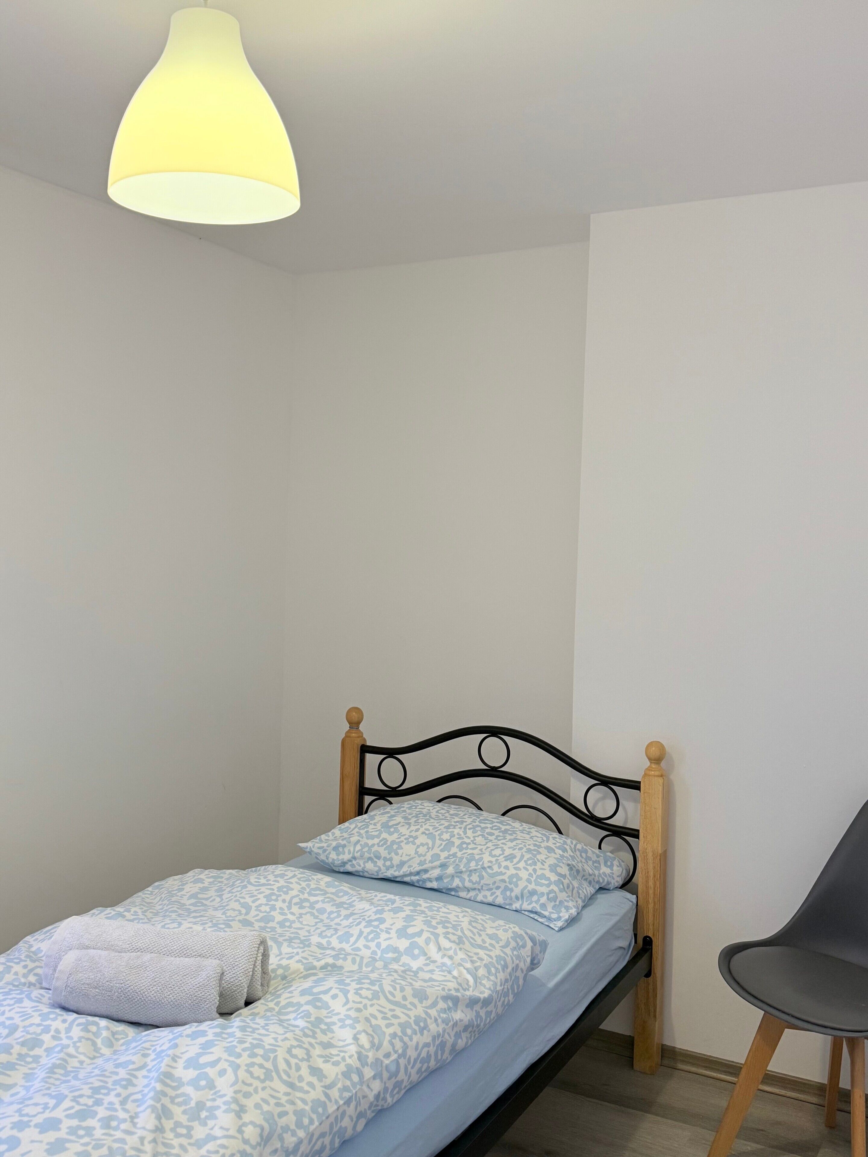 Ein Einzelbett mit Holz-Kopfteil, ein grauer Sessel und eine hängende Leuchte.