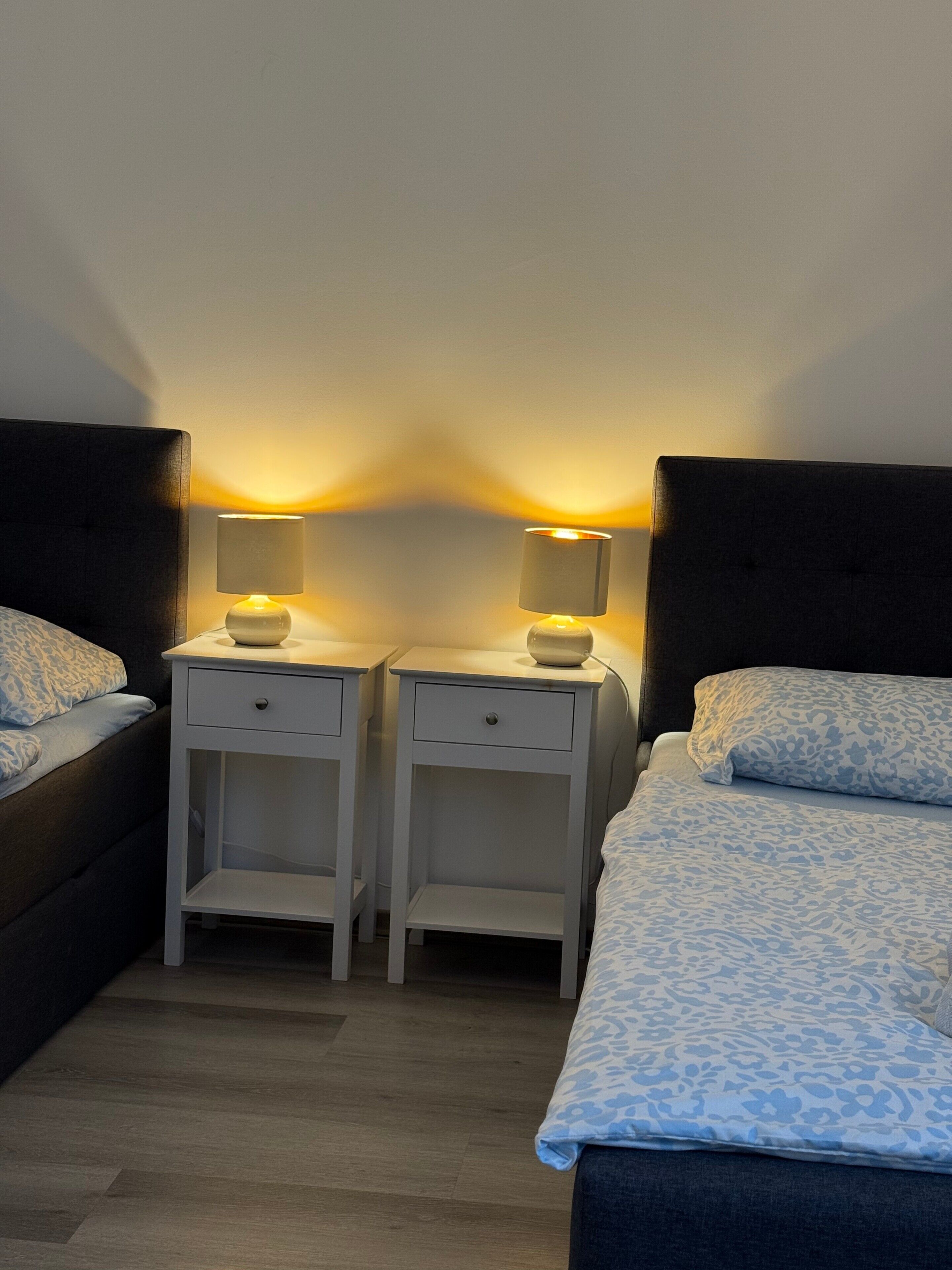 Un dormitorio con dos mesitas de noche, cada una con una lámpara, una cama con edredón estampado y suelo de madera.