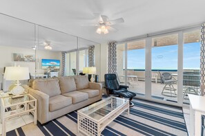 Ein Zimmer mit einer Couch, einem Couchtisch, einem Sessel, einer Lampe, einem Deckenventilator und einem großen Fenster mit Blick auf den Strand.