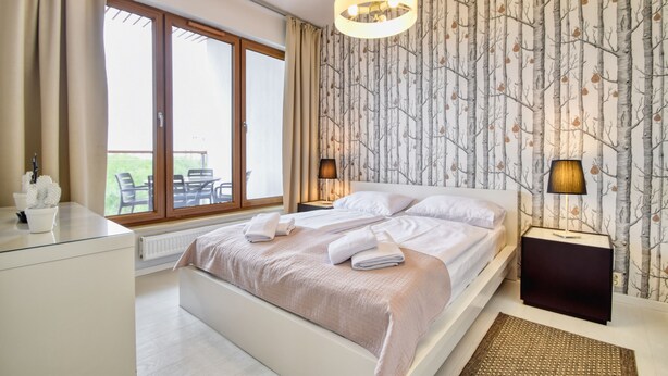 A bedroom with a large bed, a window with a view, a bedside table, and a lamp. - RentPlanet - Apartament Wschodnia (Kołobrzeg)